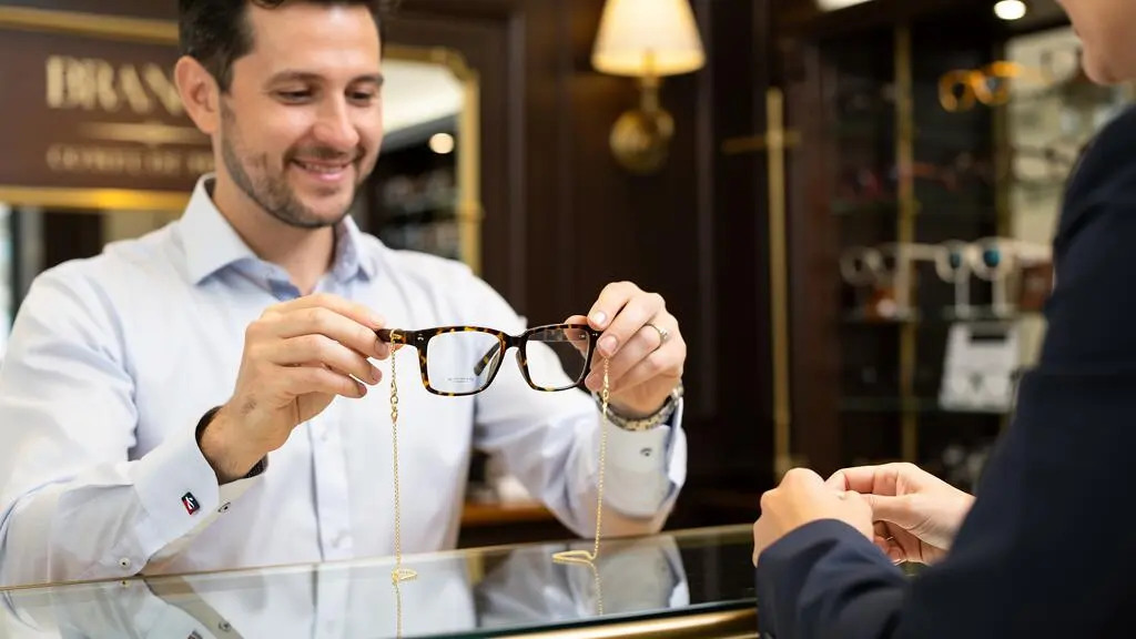 Un opticien fixe une chaîne dorée sur une monture rectangulaire lors d'un essayage en boutique.